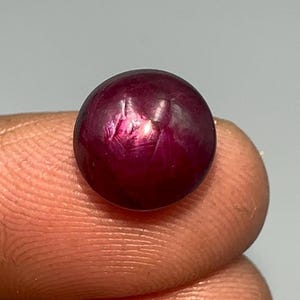 以下が含まれることがあります： 丸く磨かれた、深い赤色の宝石。 宝石は滑らかな表面を持ち、わずかな光沢があり、光を反射します。 石は2本の指の間に挟まれ、そのサイズと色を示しています。