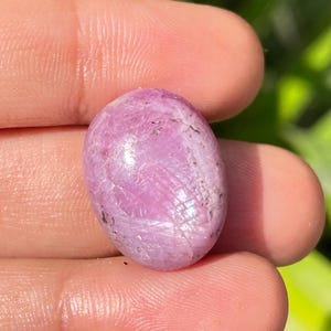 May include: A pink oval-shaped gemstone with a shimmery, iridescent finish. The stone has a rough, textured surface and is a natural, unpolished gemstone.
