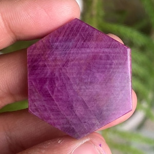May include: Hexagon-shaped, polished gemstone in a deep magenta hue. The stone has a faceted surface, catching light and revealing internal patterns. The gemstone is held between fingers, with a blurred green background.