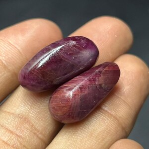 Puede incluir: Dos piedras preciosas de rubí pulidas, de forma ovalada. Las piedras preciosas son de un color rojo intenso y rico con un acabado brillante. Las piedras se sostienen en una mano, con un fondo oscuro.