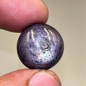 May include: A round, polished gemstone with a star effect. The gem is held between two fingers. The stone is a deep purple color with a six-rayed star pattern visible on its surface. The gem has a smooth, reflective surface.