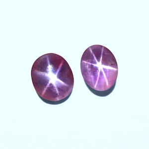 May include: Two oval-shaped gemstones exhibiting a star effect. The gems are a deep pinkish-purple hue, featuring a six-rayed star pattern emanating from the centre. The stones are set against a light blue backdrop.