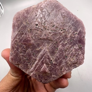 May include: A large, flat, hexagonal ruby crystal specimen with a dusty mauve and deep pink hue. The surface displays a distinct geometric triangular pattern etched into the stone, with visible dark mineral inclusions and a rough, matte texture.