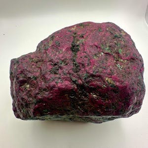 May include: A large, rough-hewn rock with a deep magenta hue, speckled with areas of dark green and lighter flecks. The surface has a textured appearance, suggesting a natural mineral formation. The overall shape is irregular, with a slightly rounded top.
