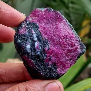 Può includere: Una pietra preziosa grezza di colore rosa e nero con una tonalità verde scuro. La pietra preziosa ha una texture grezza e naturale.