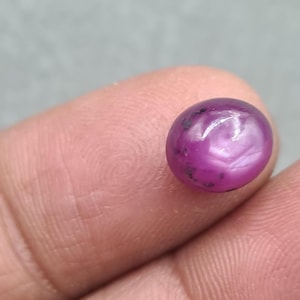 May include: A close-up of a polished, oval-shaped ruby gemstone. The gem is a deep magenta color with a subtle star effect and dark inclusions. The stone is held between two fingers.