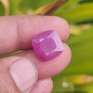 May include: A faceted, cushion-cut ruby gemstone. The gem is a vibrant pinkish-red color, with multiple facets that reflect light. The gemstone is held between fingers, with a blurred green background.
