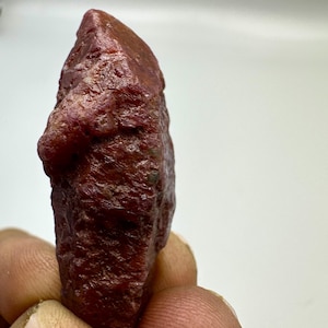 May include: A rough, uncut ruby gemstone with a deep red color. The natural stone has a textured surface and irregular shape, showcasing its raw, unpolished state. The ruby is held in a hand against a white background.