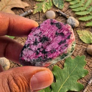 Può includere: Un esemplare minerale grezzo e non lucidato caratterizzato da una struttura cristallina color magenta vibrante e rosa intenso, con inclusioni nere sparse e macchie di materiale minerale verde pallido lungo i bordi, di forma naturale e irregolare.