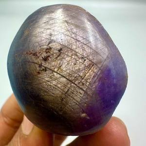 May include: A smooth, oval-shaped gemstone with a dark, purplish-gray hue. The surface is marked with fine, intersecting lines and a patch of deeper purple. The stone is held in a hand, suggesting its size and texture.