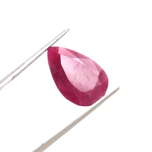 Puede incluir: Una gema de rubí facetada en forma de pera, sujeta con pinzas plateadas sobre un fondo blanco. El rubí muestra un color rojo intenso con múltiples facetas que capturan la luz. La gema tiene una forma alargada, con un extremo puntiagudo y una parte superior redondeada.