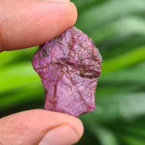 May include: A rough-cut, natural ruby gemstone. The ruby is a deep, rich pinkish-red color with visible texture and natural inclusions. The gemstone is held between two fingers against a blurred green background.