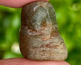 May include: A raw gemstone held between fingers, displaying a mix of brown, grey, and green hues. The stone's surface is textured, and its shape is irregular, indicating it's in its natural, unpolished state. The gemstone is likely a mineral specimen.