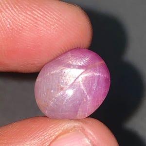 May include: A polished, oval-shaped, pink gemstone. The gem has a star-like pattern across its surface. The stone is held between two fingers, with a dark background.