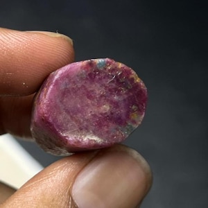 May include: A rough-cut, oval-shaped ruby gemstone. The ruby displays a deep, rich red colour with areas of lighter pink and hints of blue and yellow. The stone is held between two fingers against a dark background.