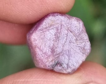 May include: A rough, uncut ruby gemstone held between fingers. The ruby has a textured surface and a light purple hue. The gemstone is irregularly shaped, with visible crystalline structures.