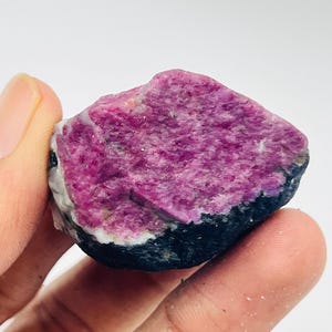 Op de afbeelding: Een ruwe, ongeslepen edelsteen met een levendig magenta interieur en een donker, zwart exterieur. De steen heeft een textuur en een kleine witte band die de twee kleuren scheidt. De edelsteen wordt in een hand gehouden.