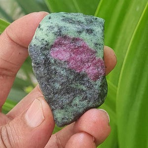 Puede incluir: Una piedra verde y negra con una inclusión rosa y roja. La piedra es rugosa y tiene un acabado natural sin pulir.