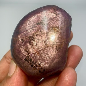 Può includere: Una gemma lucidata, di forma ovale, con una tonalità viola intenso e una superficie brillante e riflettente. La pietra ha una consistenza liscia con motivi e inclusioni interne visibili. È tenuta in mano su uno sfondo neutro.