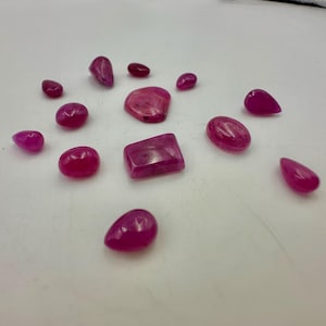 Op de afbeelding: Een verzameling gepolijste, levendige roze edelstenen in verschillende vormen, waaronder ovaal, druppelvormig en rechthoekig. De edelstenen zijn verspreid over een wit oppervlak, wat hun rijke kleur en natuurlijke variaties benadrukt.