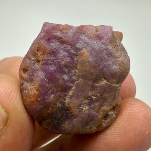 May include: A raw, uncut ruby gemstone with a deep reddish-purple hue. The natural stone has an irregular shape and a textured surface, showcasing its raw, unpolished state. The ruby displays variations in colour and subtle imperfections.