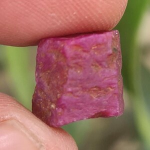 May include: A raw ruby gemstone, held between fingers. The ruby is a deep pinkish-red colour with a rough, textured surface. The gemstone has a rectangular shape with visible natural inclusions and colour variations.