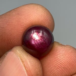 May include: A close-up of a round, polished, deep red gemstone. The gem has a star-like pattern of light reflecting within its surface. The stone is held between two fingers, showcasing its size and color.