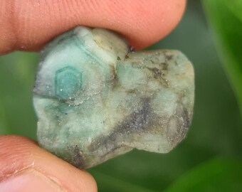 May include: A rough-cut, natural emerald gemstone. The emerald is held between two fingers and displays a mix of green and dark gray hues. The gemstone has a hexagonal pattern on its surface.