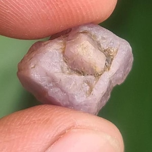 May include: A raw, unpolished gemstone with a pale pink and translucent appearance. The stone features an irregular, multifaceted surface with visible natural textures, light-colored inclusions, and a soft, cloudy crystalline structure throughout.