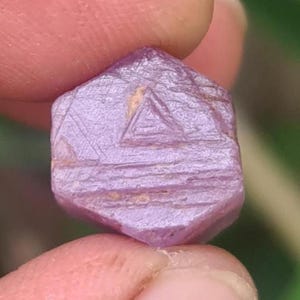 Op de afbeelding: Een ruwe, ongeslepen, zeshoekige robijn edelsteen. De robijn wordt tussen twee vingers gehouden en heeft een lichtpaarse tint met zichtbare oppervlaktestructuur en een driehoekige markering op één kant. De edelsteen is een natuurlijk mineraal.