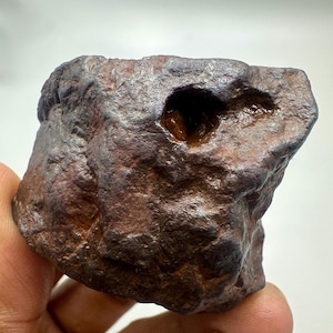 Könnte beinhalten: Ein rauer, dunkelbrauner Felsen mit einer strukturierten Oberfläche und einem großen, unregelmäßigen Loch. Der Felsen hat einen metallischen Glanz und scheint eine natürliche Formation zu sein. Die Gesamtform ist etwas rechteckig, mit abgerundeten Kanten.