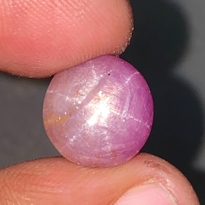 May include: A round, polished, pink gemstone held between fingers. The gem has a star-like effect across its surface, with subtle variations in color and a smooth, reflective finish. The gemstone is a natural, unmounted jewel.