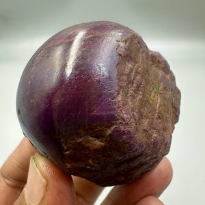 May include: A natural ruby gemstone, showing both polished and rough surfaces. The polished area is a rich, deep purple-red with a glossy finish. The unpolished side has a textured, brown appearance.
