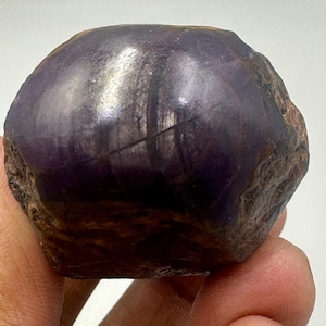 May include: A polished, dark purple gemstone with a smooth, reflective surface. The stone has a rounded shape with a rough, textured edge. The color varies from deep violet to nearly black, with subtle light reflections.