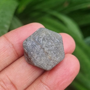 May include: A grey, hexagonal gemstone with a rough, textured surface. The stone is held in a hand, with a blurred green background. The gemstone appears to be a raw, uncut mineral.
