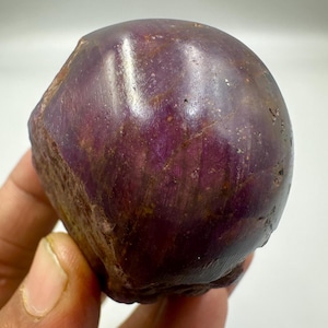 Può includere: Una gemma grezza, non tagliata, di colore viola intenso, che presenta trame e variazioni naturali. La superficie della pietra ha un aspetto lucido, con aree di tonalità più chiare e più scure. La forma generale è arrotondata, con alcuni bordi ruvidi.