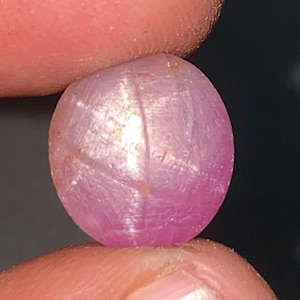 May include: A round, polished, pink gemstone with a star-like pattern across its surface. The gem is held between two fingers, showcasing its smooth, reflective texture and subtle color variations. The gem is a ruby.