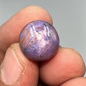 May include: A round, polished gemstone with a star effect. The gem is a mix of purple and pink hues, with a visible star pattern across its surface. The stone is held between fingers, showcasing its size and color.