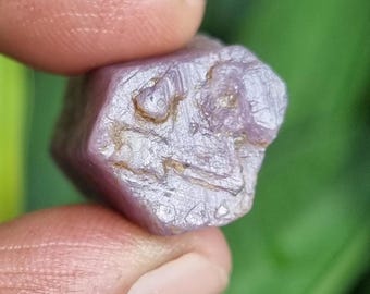 May include: A rough, uncut, light purple gemstone with a crystalline structure. The stone is held between fingers, with a blurred green background. The gemstone has a slightly irregular, faceted shape and a matte finish.