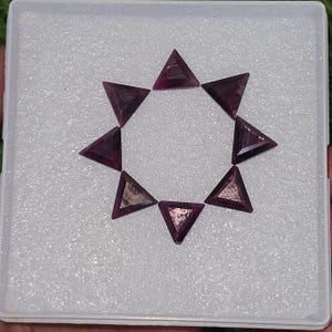 May include: A collection of eight triangular, faceted gemstones arranged in a starburst pattern. The gems are a deep, rich burgundy color and are displayed in a white, textured tray. The facets catch the light, creating a sparkling effect.
