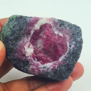 May include: A raw, unpolished mineral specimen featuring a central cluster of vibrant magenta-pink ruby crystals surrounded by a ring of white quartz, all embedded within a dark, textured grey and green zoisite matrix with a rough, natural surface.