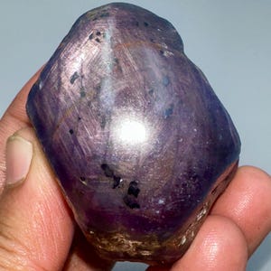 May include: A rough, uncut gemstone in a deep purple colour, held in a hand. The stone has a textured surface with streaks of lighter colour and small black inclusions. The overall shape is irregular and organic.