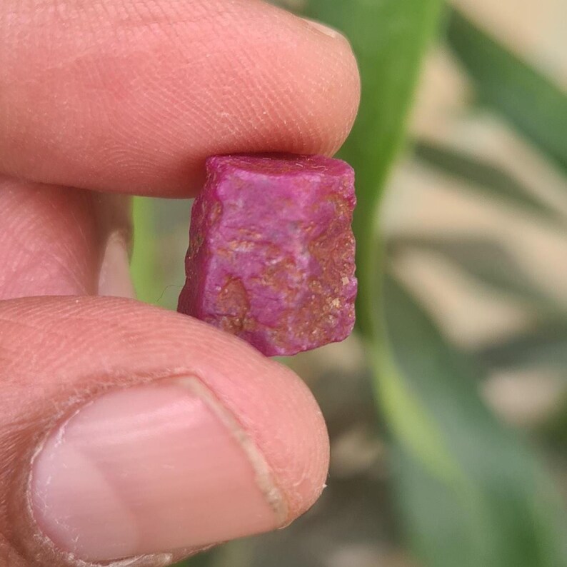May include: A raw ruby gemstone, held between fingers. The ruby is a deep pinkish-red colour with a rough, textured surface. The gemstone is a small, rectangular shape, showcasing its natural, unpolished state. The background is blurred.