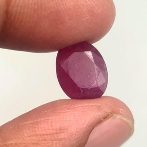 May include: A deep magenta-colored oval gemstone with a faceted surface. The stone features a polished finish and multiple geometric cuts that reflect light across its translucent, saturated surface, showcasing a vibrant and rich pinkish-red hue.