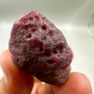 May include: A rough, uncut ruby gemstone. The ruby is a deep, rich red color with a textured surface and several small holes. The gemstone is held in a person's hand.