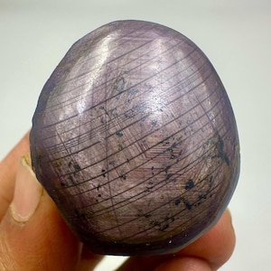 May include: A polished, oval-shaped gemstone with a deep purple hue. The surface features a pattern of intersecting lines, creating a textured appearance. The stone is held in a hand, showcasing its size and shape.