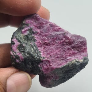 Peut inclure: Un spécimen minéral brut et non poli présentant une section cristalline magenta-rose vif contrastant avec une matrice rocheuse noire opaque et sombre. La pierre a une forme irrégulière et dentelée avec une surface rugueuse.