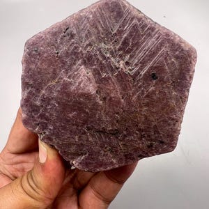 May include: A large, raw, hexagonal-shaped ruby crystal with a deep purplish-red hue. The surface displays a rough, matte texture with visible striations, natural mineral inclusions, and a geometric, flat-faced crystalline structure.