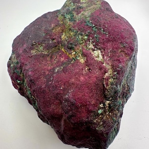 Pode incluir: Uma grande rocha de forma irregular com um tom magenta profundo, salpicada com manchas verdes e douradas. A superfície tem uma aparência áspera e texturizada, sugerindo uma formação mineral natural. A forma da rocha é sólida e substancial.