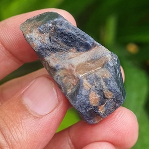 May include: A raw, natural sapphire gemstone. The sapphire displays a deep blue color with areas of brown and gray. The stone is held in a hand, showcasing its rough, unpolished texture and natural beauty. The sapphire is a unique, one-of-a-kind item.